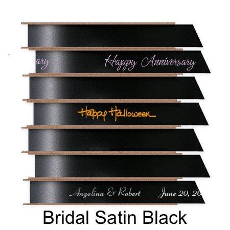 A stack of seven spools of shiny black satin ribbon with custom messages printed on them in different colors.
