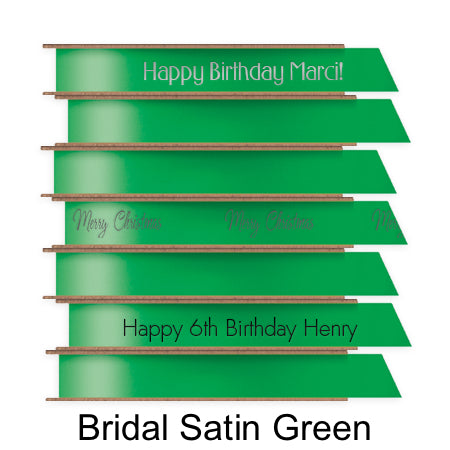 A stack of seven spools of shiny green satin ribbon with custom messages printed on them in different colors.