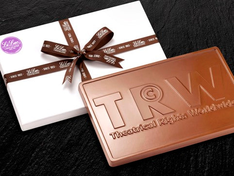 A large 1 pound chocolate bar has a business logo T R W embossed onto it. It leans on a white gift box tied with a brown Li-Lac Chocolates branded ribbon.