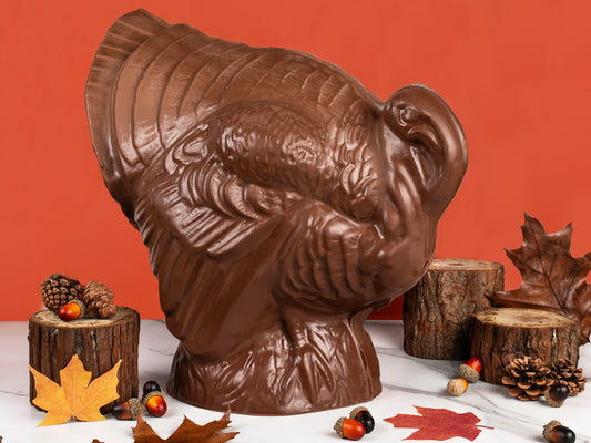 A giant chocolate turkey standing on a white marble table with tree branch slices, acorn and pinecone decorations.