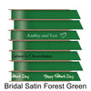 A stack of seven spools of shiny forest green satin ribbon with custom messages printed on them in different colors. 