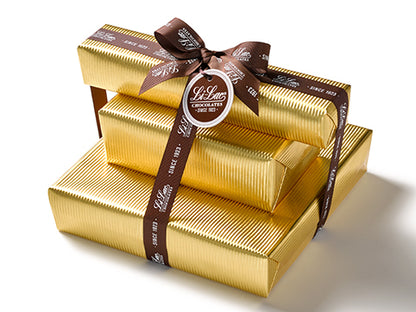 Three boxes of chocolate have been beautifully gift wrapped in gold foil paper. They are stacked on top of each other and tied together with a dark brown Li-Lac Chocolates logo printed ribbon with a round gift tag.