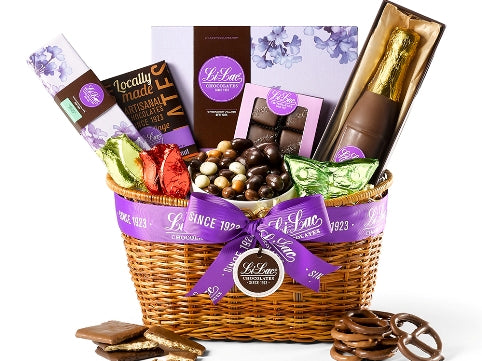 A low, oval wicker basket is tied with a purple Li-Lac Chocolates ribbon. The basket holds one home assortment chocolate box, box of French mints, one boxed chocolate bar, a bag of chocolate espresso beans, two chocolate tulips and one chocolate butterfly wrapped in colorful foil, salted caramels in a purple window box, a 7” chocolate champagne bottle.