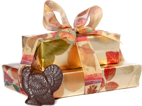 Two boxes of chocolate have been beautifully gift wrapped in gold foil paper with an autumn leaf motif. They are stacked on top of each other and tied together with a gold and autumn leaf printed ribbon. A small chocolate turkey pop sits next to the boxes.