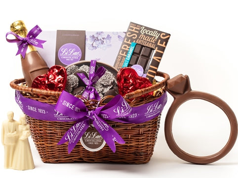 A low, oval wicker basket is tied with a purple Li-Lac Chocolates ribbon. The basket holds one chocolate diamond Ring, on chocolate Bride & Groom, a chocolate 7" champagne bottle, two chocolate hearts, one home assortment chocolate gift box, a box of chocolate nonpareils, one boxed chocolate bar.